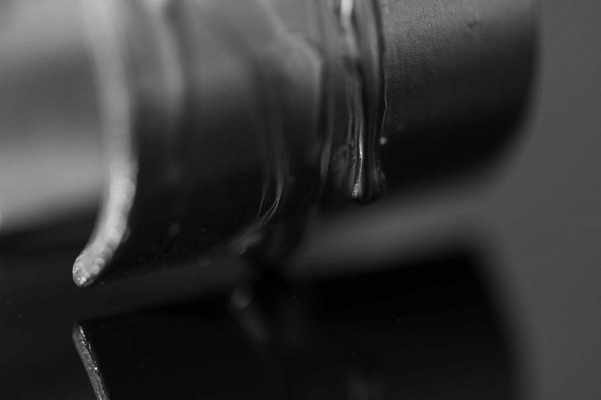 A thick dark liquid dripping from a cylindrical object onto a reflective surface